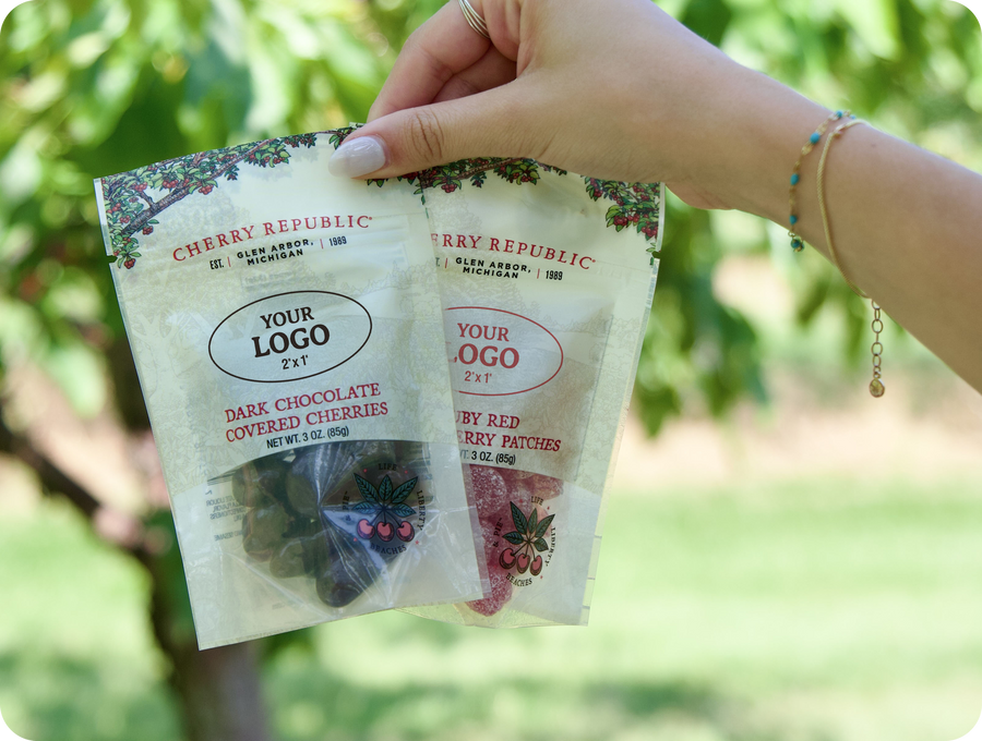 A hand holding two custom-labeled Cherry Republic 3 oz pouches in a cherry orchard